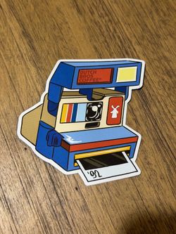 Dutch Bros Blue and Red Polaroid-Style Camera Sticker. Vinyl sticker depicting a stylized Polaroid camera with Dutch Bros branding. Bold graphic color