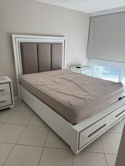 White wood bed frame with a tall upholstered headboard featuring three vertical panels and nailhead trim. Includes under-bed storage drawers at the fo