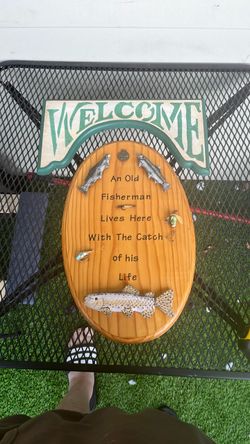 is a rustic wooden fishing-themed wall plaque that reads "An Old Fisherman Lives Here With The Catch of His Life