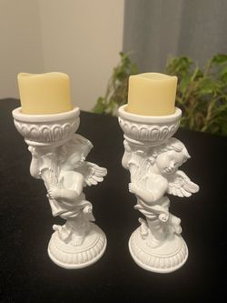 Elegant pair of white resin cherub angel candle holders. Each holder is paired with a cream-colored battery-operated LED pillar candle. The set has a