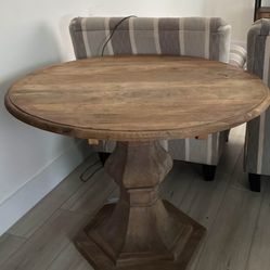 The round wooden table has a solid pedestal base, which is a design usually found in higher-quality, heavy, farmhouse or restoration-style furniture. 