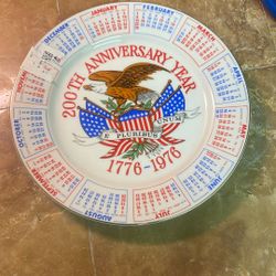 This item is a vintage 1975 Spencer Gifts calendar plate commemorating the United States Bicentennial (1776–1976).     
