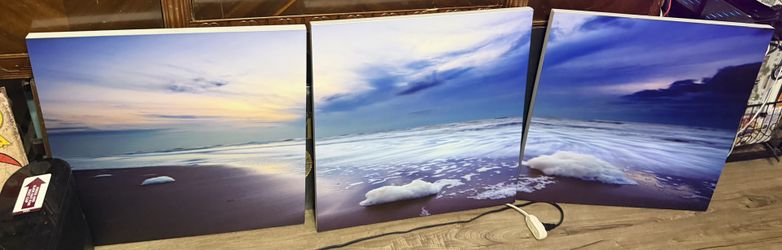 19.5 x 19.:/Preowned three-piece canvas wall art set, wide-angle photograph of a serene beach at sunset or sunrise. The scene captures foamy waves lap