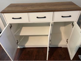 Sturdy farmhouse sideboard has white finish with contrasting wood-look top. Cabinet features three top drawers with black metal handles and three lowe