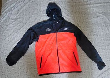 New Balance lightweight hooded windbreaker featuring a vibrant orange and dark navy color block with a reflective logo on the chest.