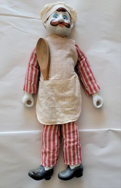 vintage porcelain chef doll featuring a painted mustache, a fabric chef's hat, red-and-white striped clothing, a small cloth apron with a wooden spoon