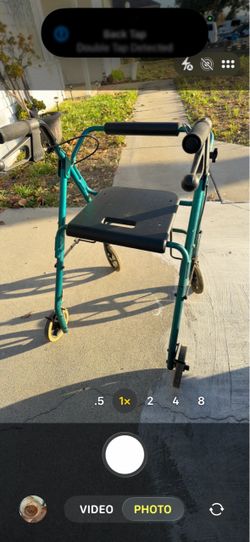 The image shows a teal four-wheel rolling walker (rollator) with a black padded seat, hand brakes, and black handlebar grips, closely resembling a Gra