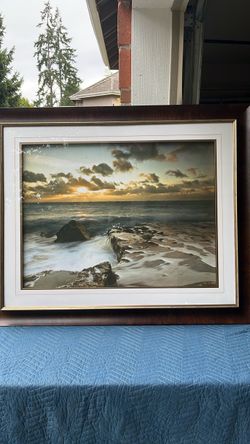 This framed coastal sunset seascape painting features expressive warm golden tones, crashing ocean waves against rocky cliffs, and a rich dark wood fr