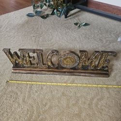 decorative wooden "Welcome" sign with a gold-tone finish and ornate detailing. 