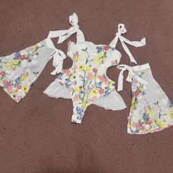 White one-piece with colorful floral embroidery. Features tie straps and sheer skirt panels. Bright yellow and pink flowers with green stems. Playful 