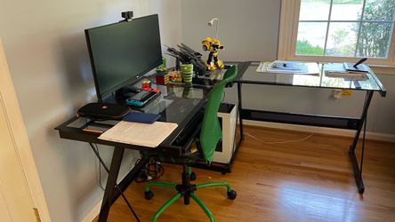 This setup features a home office or gaming workspace built around an L-shaped glass-top desk and a compact green swivel chair.