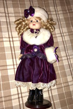 Beautiful porcelain doll with blond hair, purple dress with matching hat. Comes with doll stand.