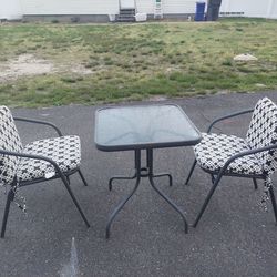 Out.\n Door metal table with glass top and 2 metal chairs with cushions.